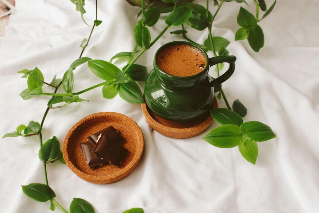 A green ceramic mug of coffee with chocolate pieces and lush green leaves on a white background.