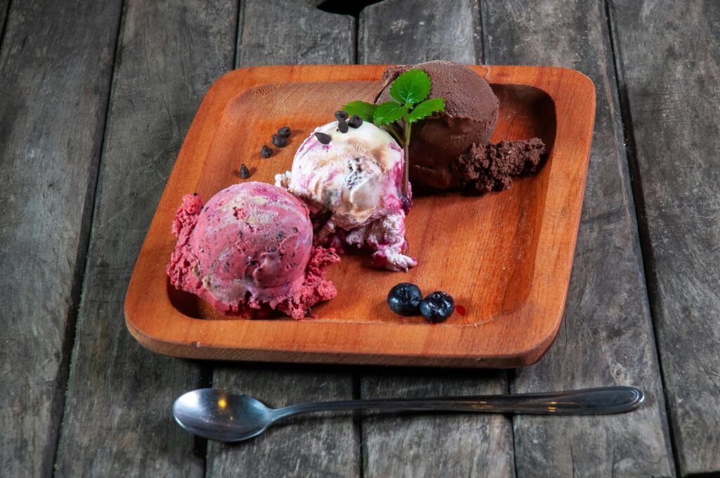 Three artisanal ice cream scoops on a wooden plate with chocolate chips and blueberries.